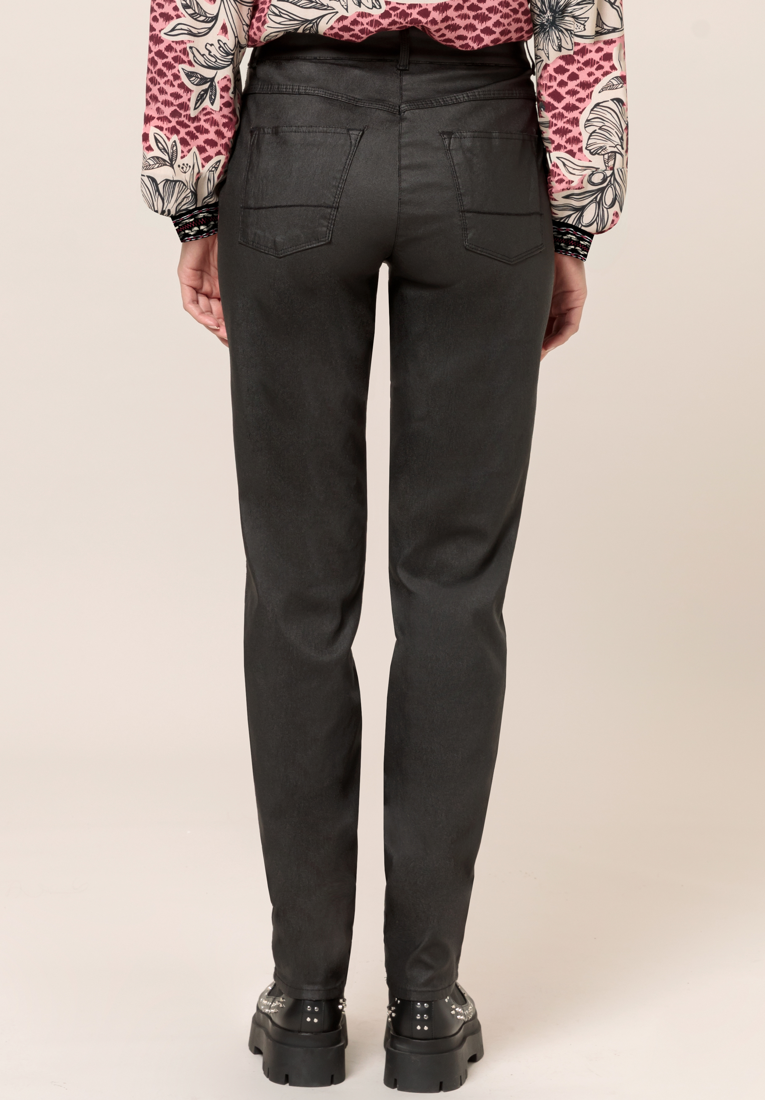 Person in schwarzen Slim-Fit-Jeans, schwarzen klobigen Schuhen und einer gemusterten Bluse mit rosa und weißen Blumenmustern, die mit dem Rücken zur Kamera vor einem schlichten hellen Hintergrund steht.