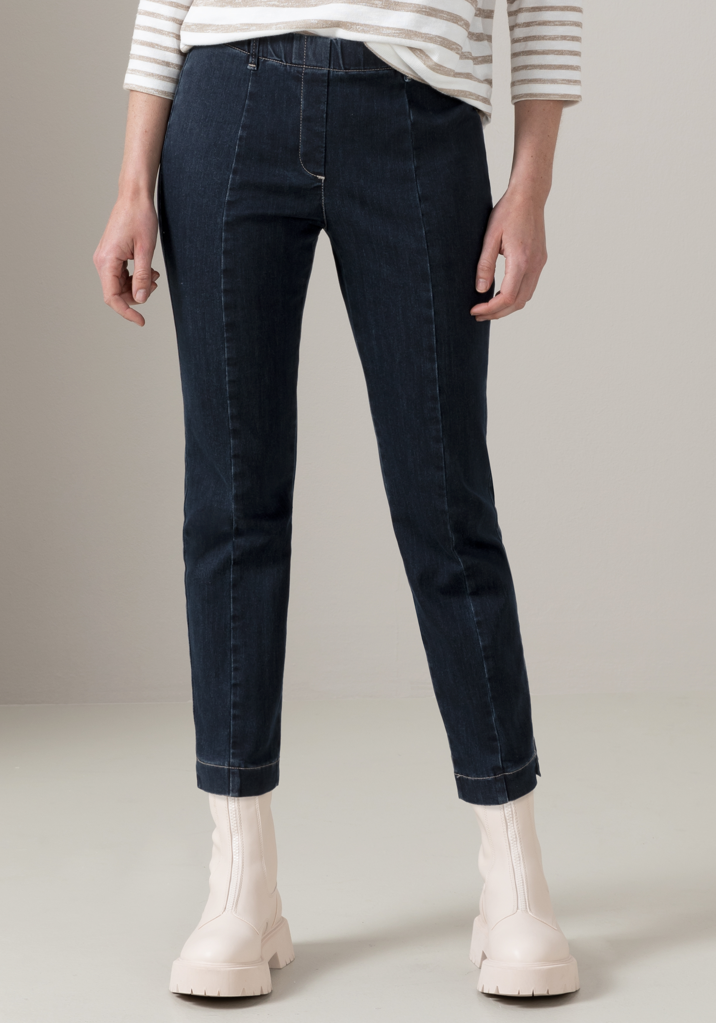 Eine Person, die dunkelblaue Jeans, ein weiß-beige gestreiftes Langarmhemd und helle, klobige Stiefel trägt, steht vor einem neutralen Hintergrund. Das Foto ist ab den Schultern nach unten beschnitten.