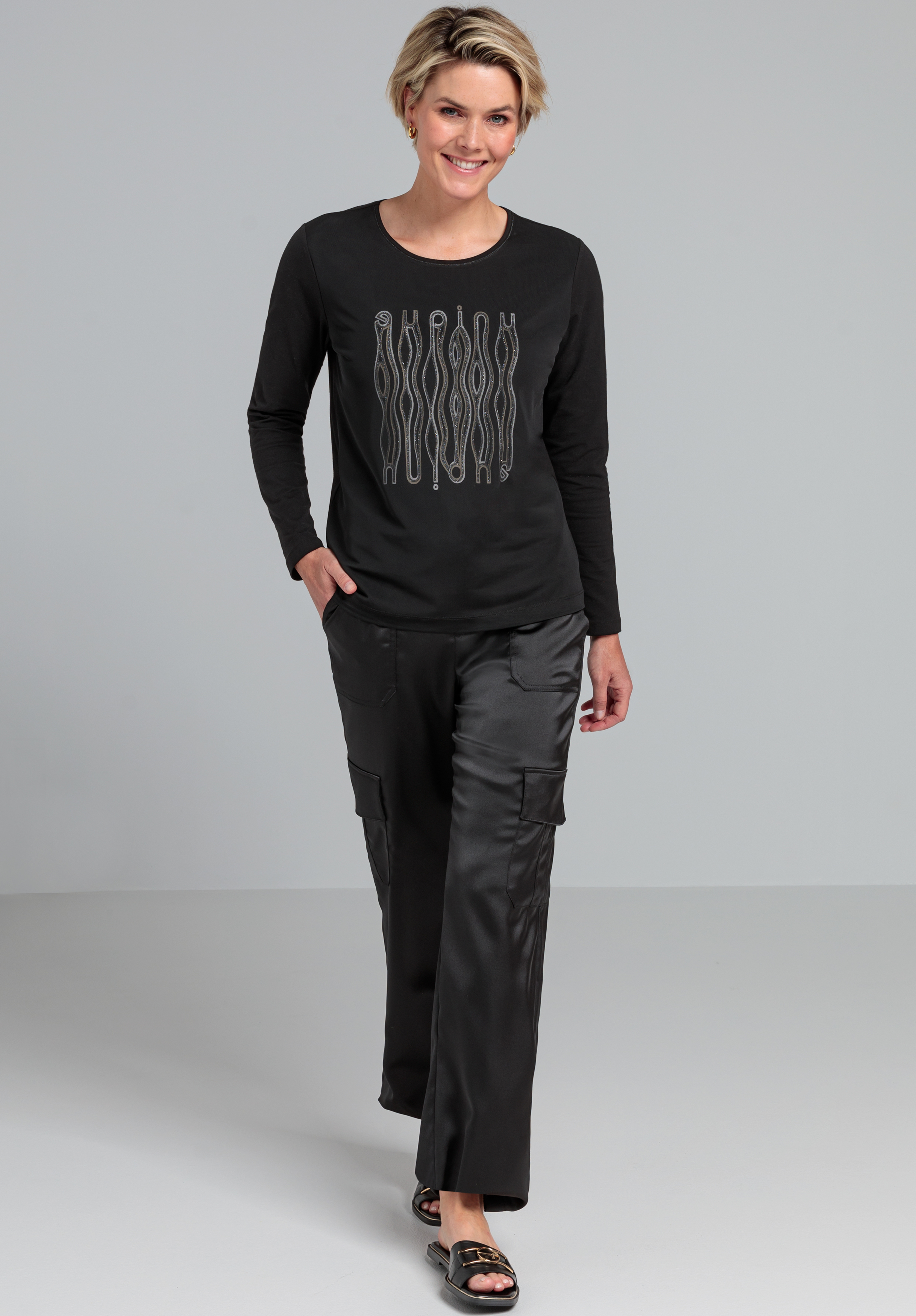 Eine lächelnde Frau mit kurzen blonden Haaren trägt ein schwarzes Langarmshirt mit Wellenlinienmuster und eine schwarze Cargohose. Sie geht mit einer Hand in der Tasche vor einem schlichten grauen Hintergrund vorwärts.