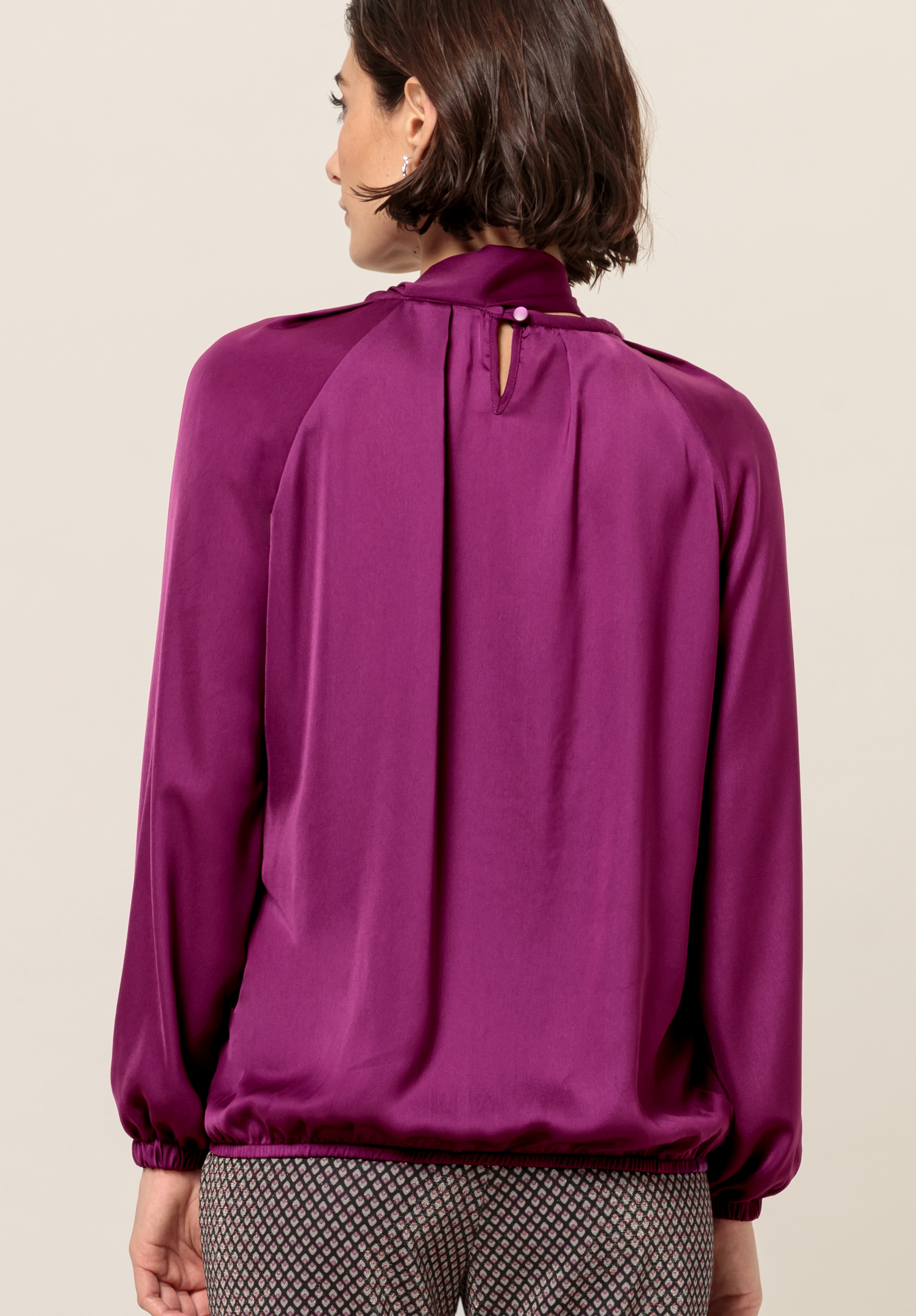 Von hinten ist eine Frau mit kurzen braunen Haaren zu sehen, die eine langärmlige, glänzende lila Bluse mit einem geknöpften Schlüsselloch am Hals trägt, dazu eine gemusterte Hose.