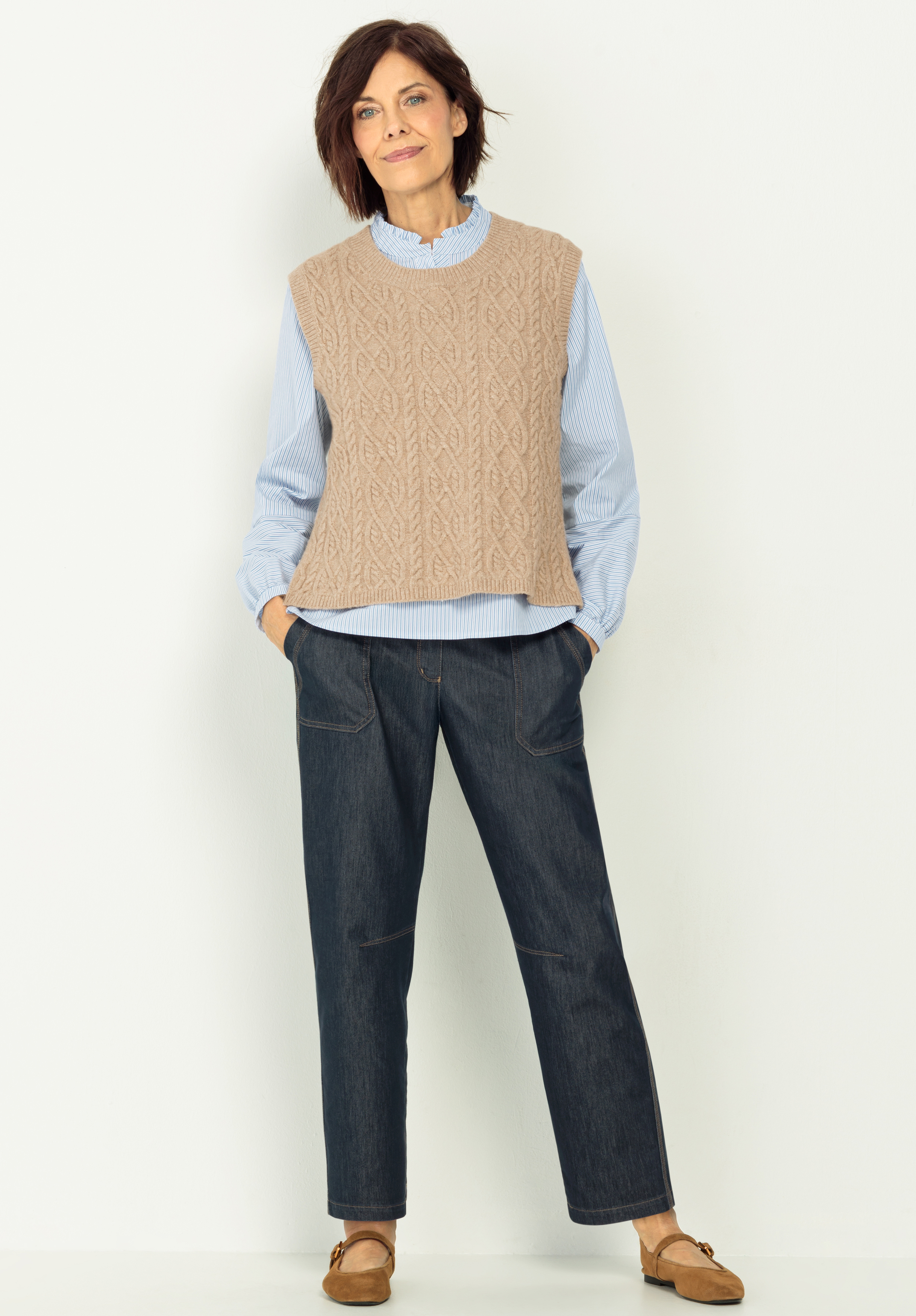 Eine Frau steht vor einer hellen Wand und trägt eine beigefarbene Strickweste über einer hellblauen Bluse, dunkelblaue Jeans und braune Halbschuhe. Sie hat kurzes braunes Haar und lächelt mit den Händen in den Taschen.