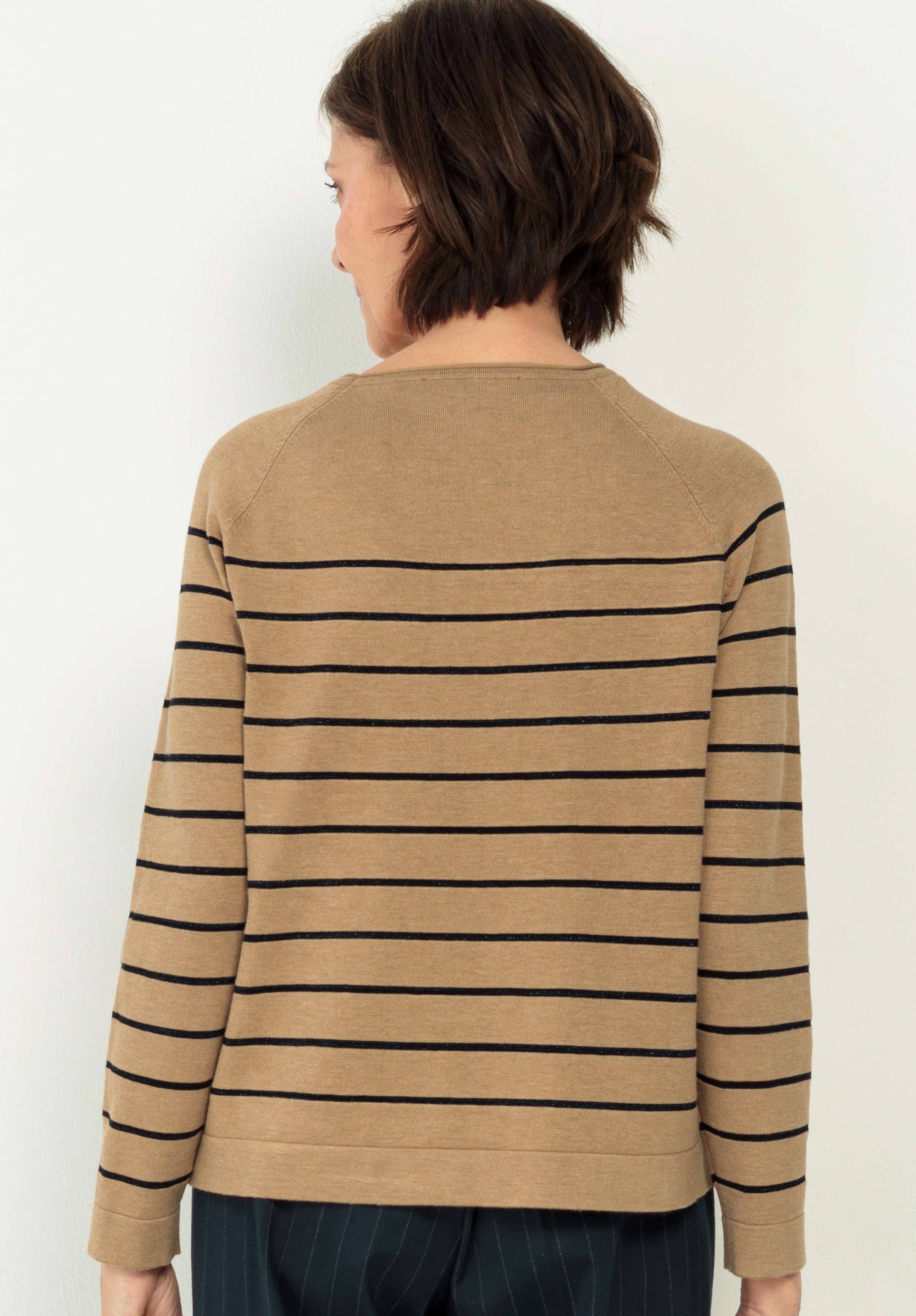 Eine Person mit kurzen braunen Haaren wird von hinten gezeigt. Sie trägt ein hellbraunes langärmeliges Oberteil mit schwarzen Querstreifen und eine dunkle Hose. Der Hintergrund ist einfarbig und hell.