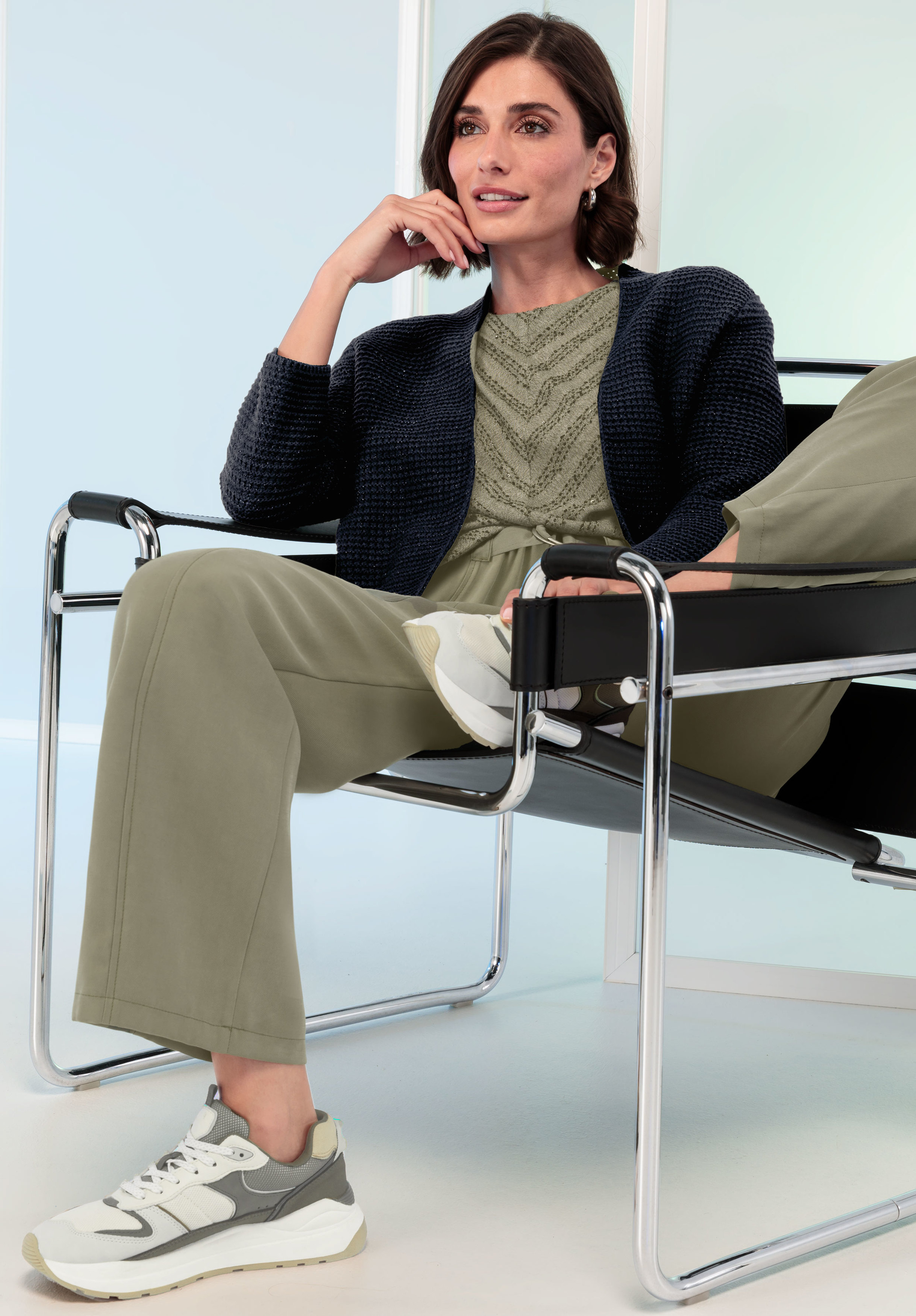 Frau sitzt lässig in einem modernen schwarzen Stuhl, trägt olivgrüne Hosen, ein gemustertes grünes Oberteil, eine strukturierte marineblaue Strickjacke und weiße Turnschuhe.