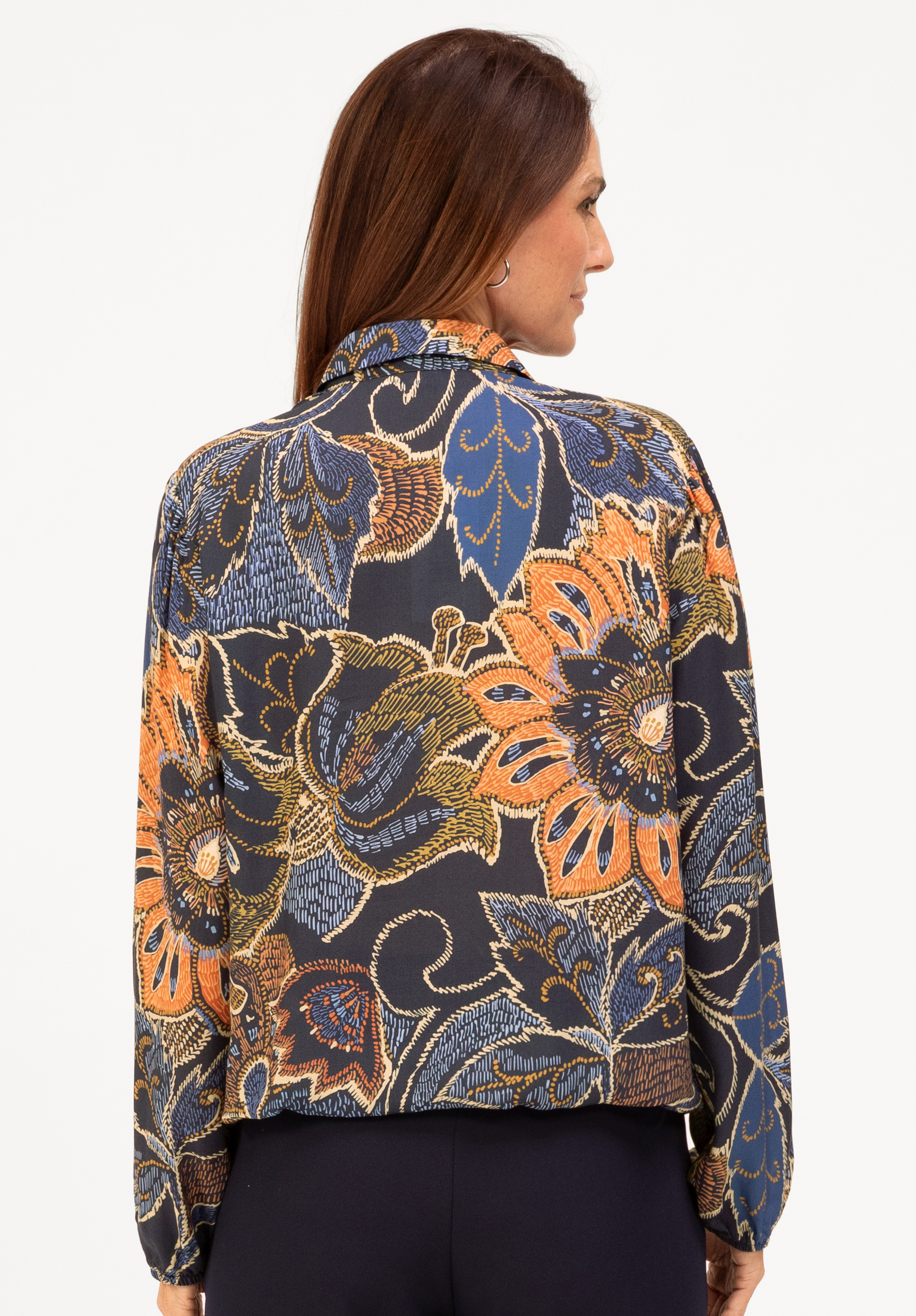 Eine Frau mit langen braunen Haaren trägt eine dunkle Bluse mit einem kräftigen, bunten Blumenmuster in Orange-, Blau- und Beigetönen. Sie blickt von sich weg und zeigt die Rückseite der Bluse und der dunklen Hose.